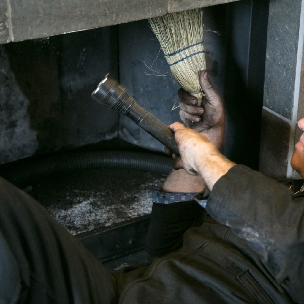 person holding a flashlight and a small broom, crouched down, looking up into a dirty fireplace to inspect it